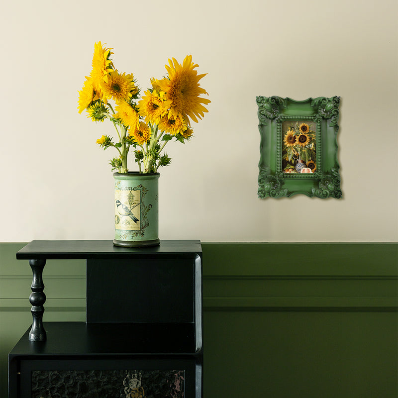 A vintage picture frame with hand-carved floral details in dark green, displayed around a sunflower painting.