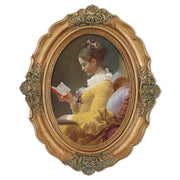 An oval-shaped, gold-colored picture frame with embossed details, containing a framed artwork of a person reading.