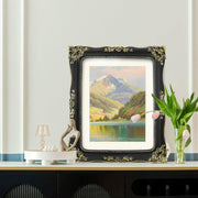 A vintage-inspired black picture frame displays a landscape painting on a shelf with decorative items.