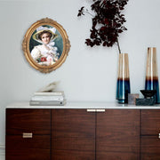 An oval-shaped, gold-colored picture frame with embossed details, containing a framed artwork of a person reading.
