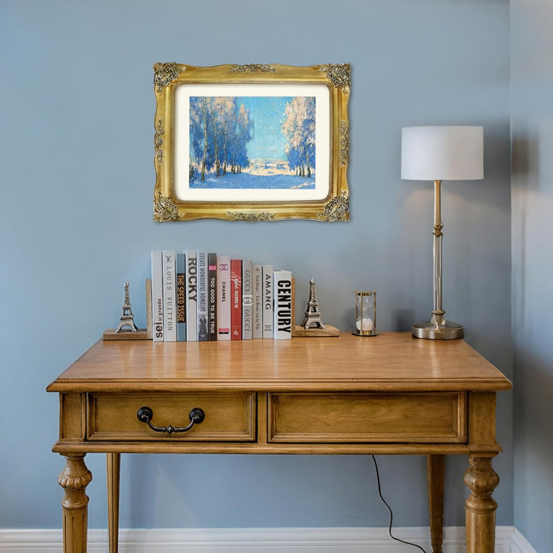 Wooden desk with books, a lamp, and a vintage gold picture frame with painting against a blue wall.