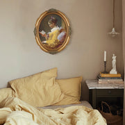 An oval-shaped, gold-colored picture frame with embossed details, containing a framed artwork of a person reading.