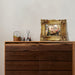 Wooden dresser with decorative items including a framed painting on a plain wall.