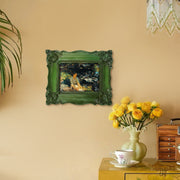 Decorative green frame with a painting on a beige wall, next to a vase of yellow flowers and a cabinet.