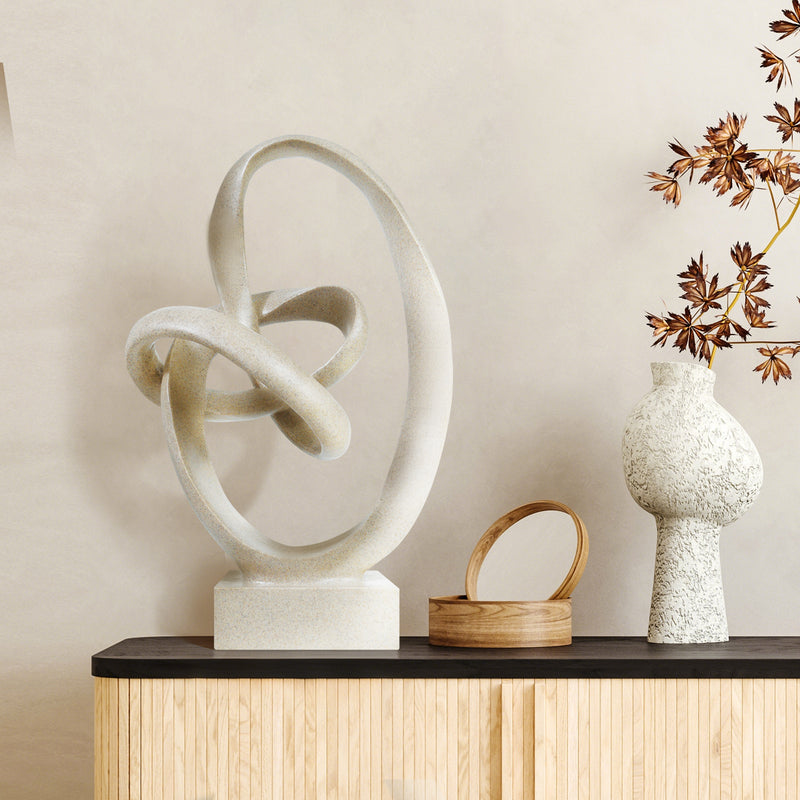 Abstract sculpture on a wooden shelf with a vase and decorative plant against a beige wall.