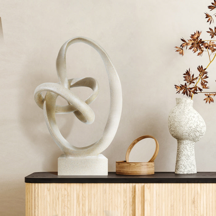 Abstract sculpture on a wooden shelf with a vase and decorative plant against a beige wall.