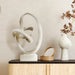 Abstract sculpture on a wooden shelf with a vase and decorative plant against a beige wall.