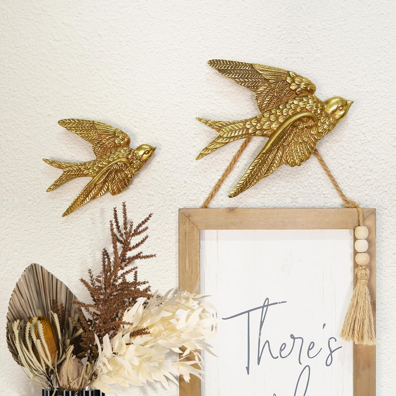 Decorative wall art with gold birds, dried plants, and a wooden frame on a textured white background.