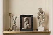 Decorative shelf with a framed portrait, diffuser, and sculpture against a beige wall.