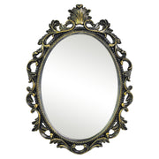 An oval mirror with a scroll design in an antique black with gold finish, reflecting a well-lit interior room.
