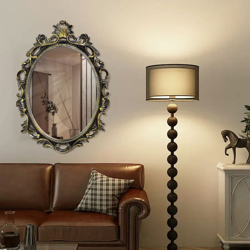 An oval mirror with a scroll design in an antique black with gold finish, reflecting a well-lit interior room.