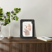 Framed abstract art on a wooden surface with a vase of greenery and a candle.