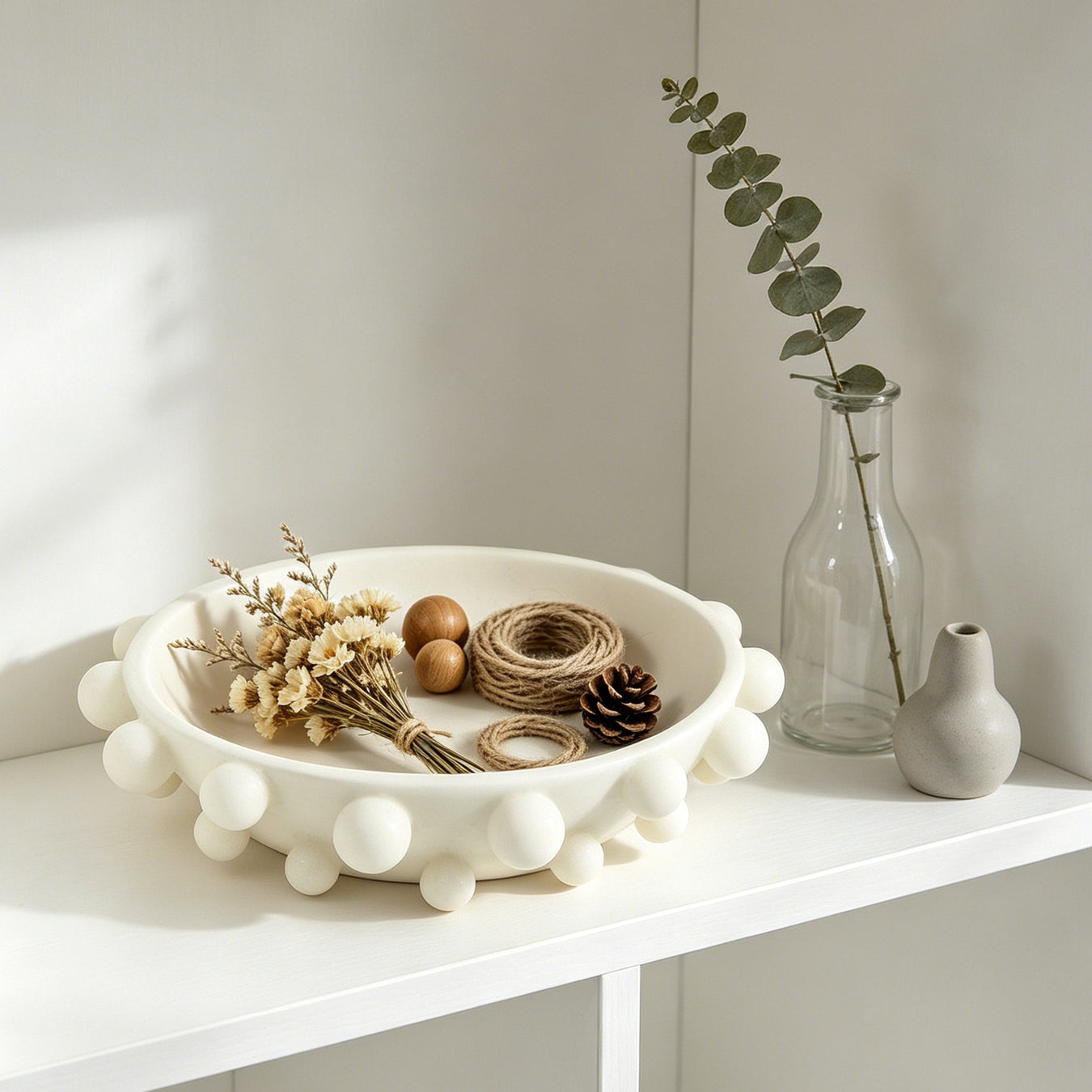 Decorative items on a white shelf including a bowl with flowers, a vase with greenery, and a small bottle.