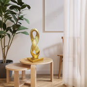 Gold abstract sculpture on a wooden table with a plant and curtain in the background.