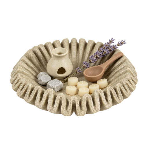 A beige textured ceramic spa tray with a wavy rim, styled with a small cream-colored diffuser, a wooden spoon, lavender sprigs, and small stones on a plain white background.