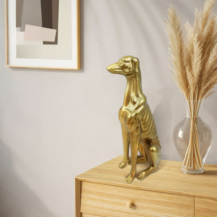 Gold dog statue on a wooden surface with a vase of pampas grass and framed picture in the background.