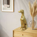 Gold dog statue on a wooden surface with a vase of pampas grass and framed picture in the background.