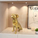 Gold dog statue on a shelf with a framed picture and a plant in the background.
