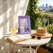 Table with book, lemonade, framed picture, sunglasses, and straw hat on a balcony with cityscape view.