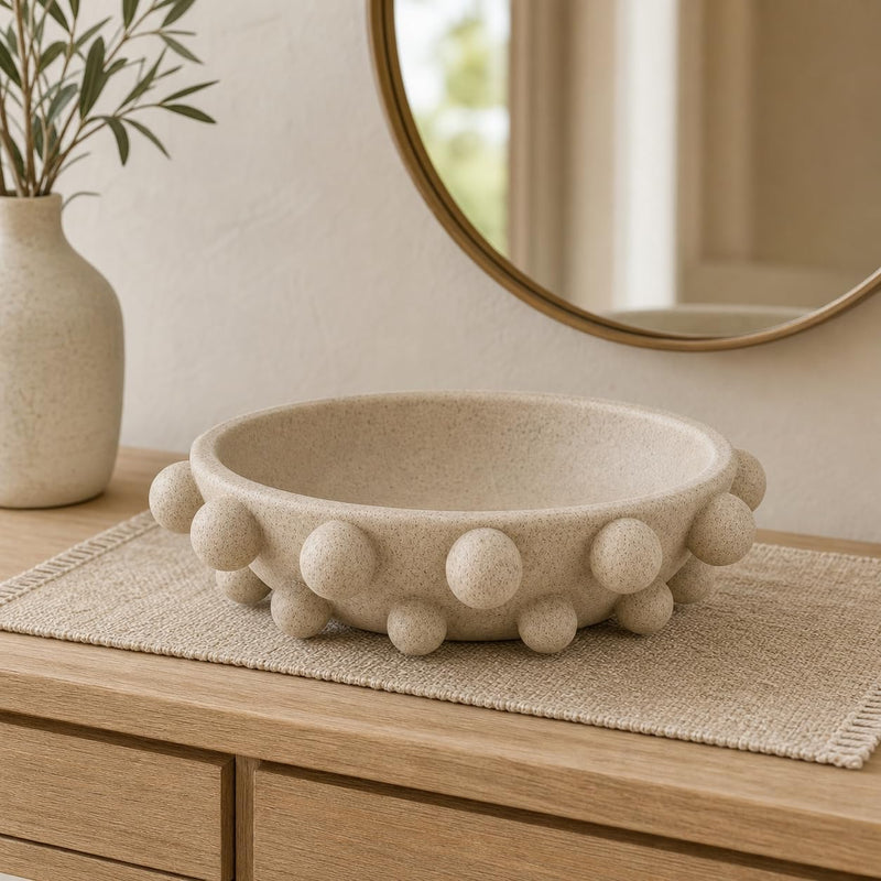 A round beige stone bowl with raised pom-pom details around the rim, styled on a woven placemat on a wooden dresser next to a vase with greenery and a round mirror.