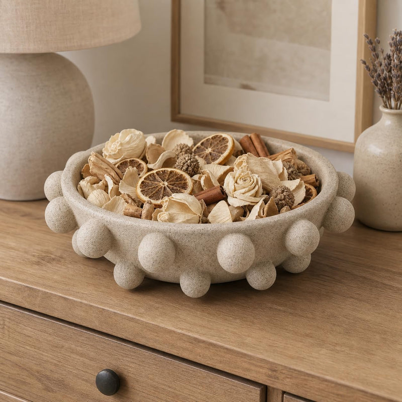 A round beige textured bowl with raised bubble-like edges, filled with dried flowers, citrus slices, cinnamon sticks, and other decorative botanicals, styled on a wooden cabinet in a home interior.