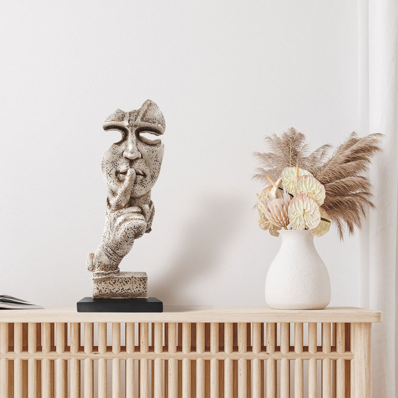 A sculpture of a human figure with an index finger to their lips, indicating silence or quiet. The figure appears to be made of a textured, possibly stone or concrete-like material, and is placed on a dark base.