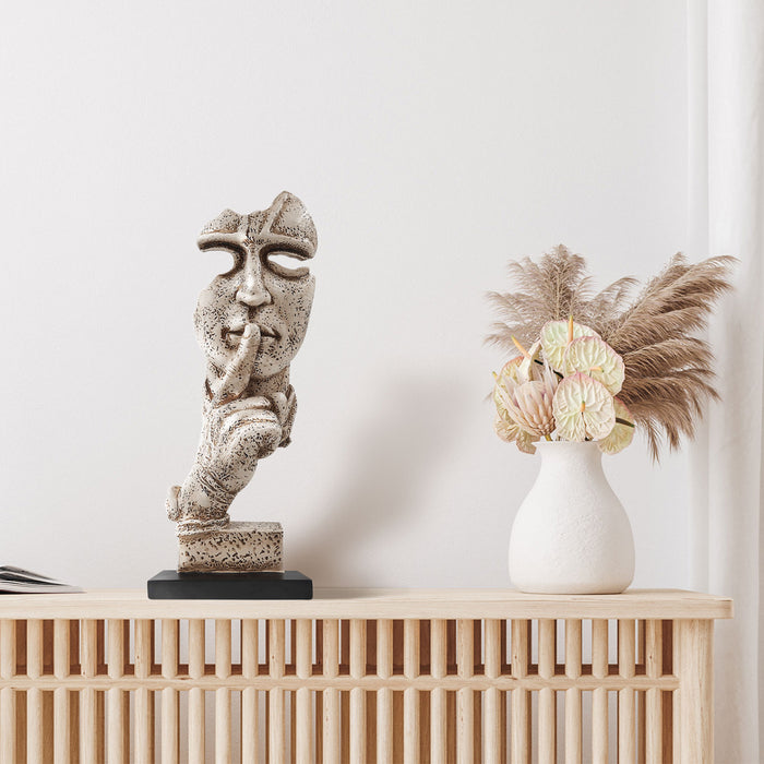 A sculpture of a human figure with an index finger to their lips, indicating silence or quiet. The figure appears to be made of a textured, possibly stone or concrete-like material, and is placed on a dark base.