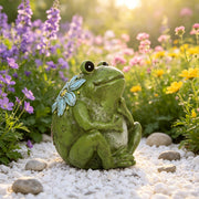 Green frog figurine with floral decorations in a garden setting with flowers and pebbles.