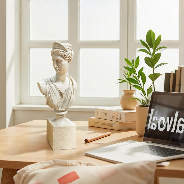 The Diana of Versailles sculptural bust stands on the desk with a laptop, books, a plant, and a statue in a bright room.