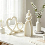 A set of minimalist white ceramic decorative home items arranged on a light marble tabletop, including two heart-shaped sculptures, a stylized couple figure, a small vase with eucalyptus stems, and a reed diffuser, photographed in a softly lit interior setting.