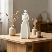 A minimalist white ceramic figurine of two abstractly joined figures stands on a light wood table, styled with a beige ceramic vase of dried grasses, a reed diffuser, and a small cup in a warm, softly blurred interior setting.