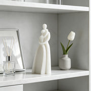 A minimalist white figurative sculpture of a couple embracing, displayed on a white shelf next to a glass reed diffuser and a small vase with a white tulip.