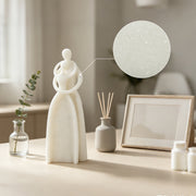 A minimalist white ceramic figurine of a woman with interlocking arms stands on a tabletop beside a small glass vase with a green sprig, a gray reed diffuser, and a blank picture frame. A circular inset close-up highlights the speckled texture of the white ceramic surface.