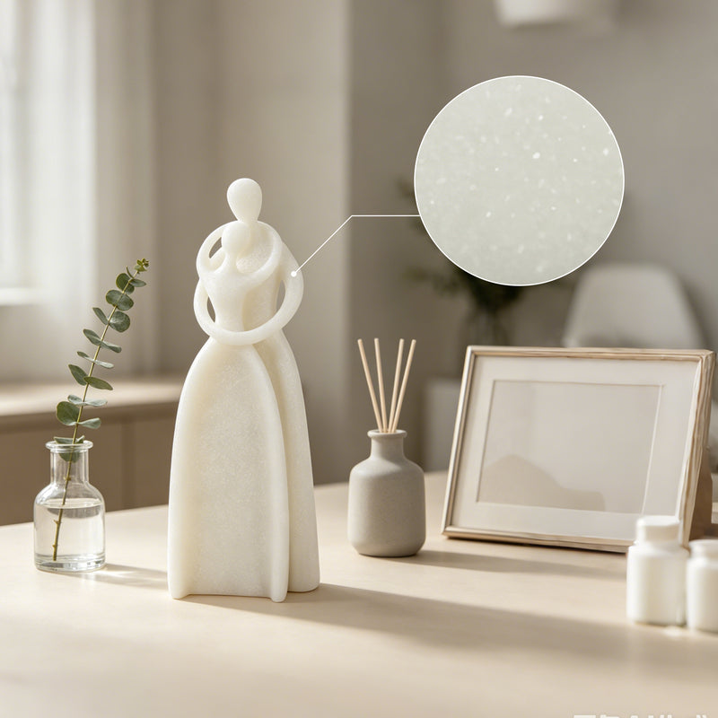 A minimalist white ceramic figurine of a woman with interlocking arms stands on a tabletop beside a small glass vase with a green sprig, a gray reed diffuser, and a blank picture frame. A circular inset close-up highlights the speckled texture of the white ceramic surface.