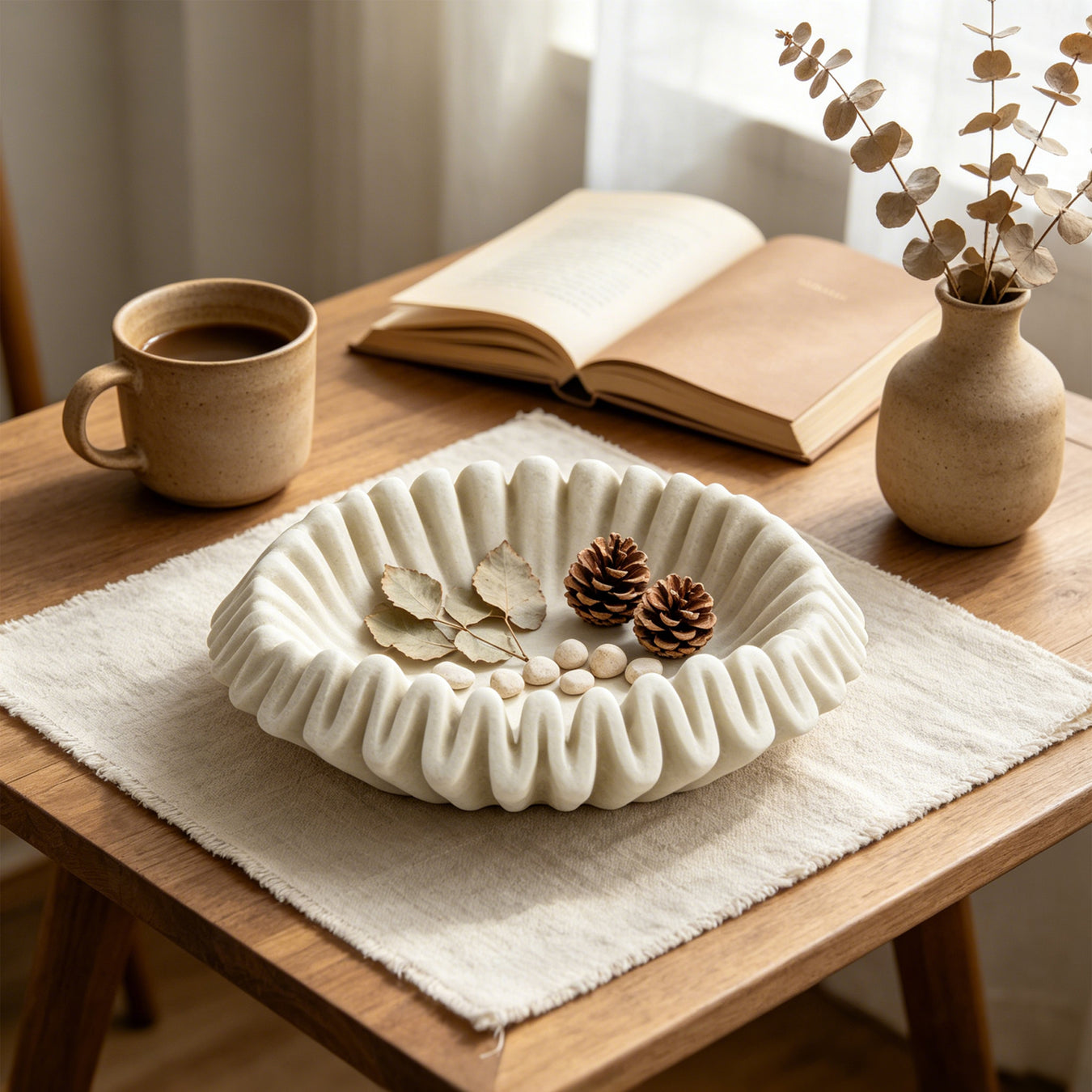 A cream-colored, scalloped ceramic bowl filled with dried leaves, pinecones, and small beads, styled on a wooden table with a mug, an open book, and a small vase.