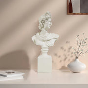White classical Apollo bust sculpture on a beige surface with a vase and books in the background.