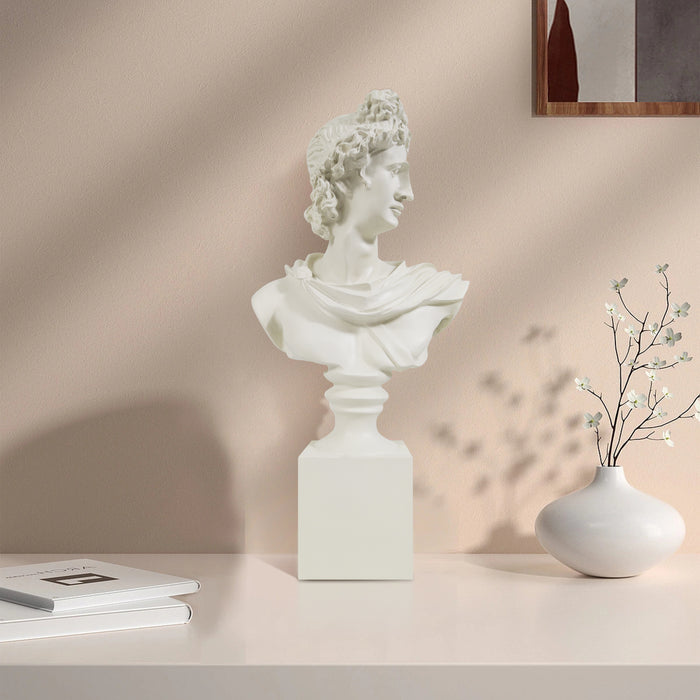 White classical Apollo bust sculpture on a beige surface with a vase and books in the background.