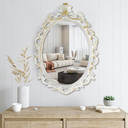 An oval mirror with a scroll design in an white with gold finish, reflecting a well-lit interior room.