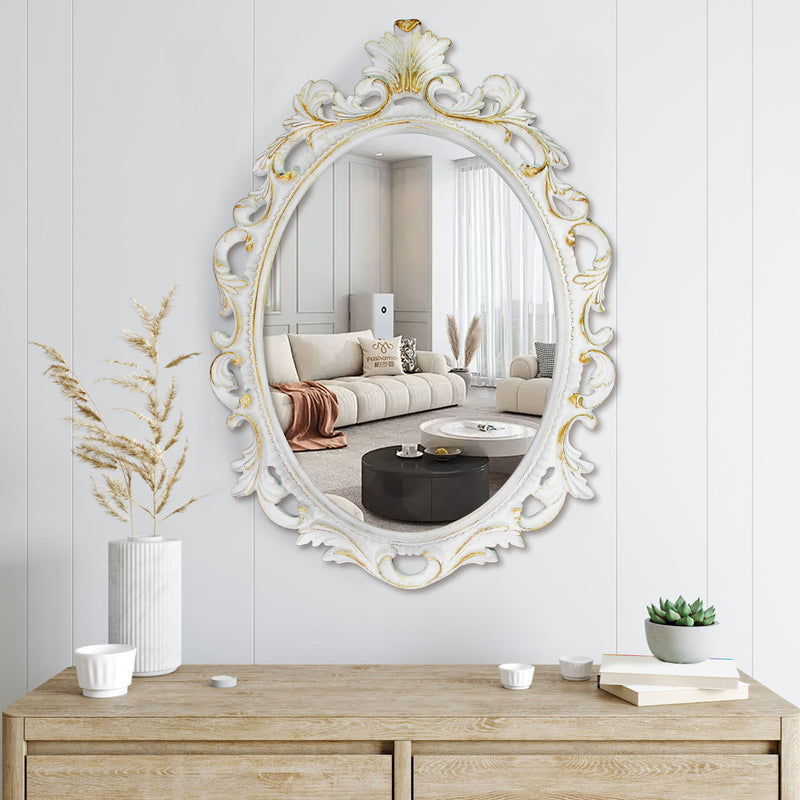 An oval mirror with a scroll design in an white with gold finish, reflecting a well-lit interior room.