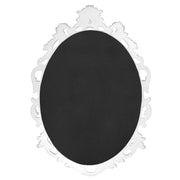 An oval mirror with a scroll design in an white with gold finish, reflecting a well-lit interior room.