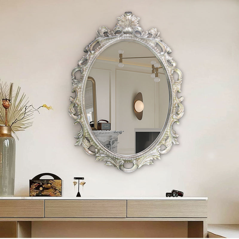 An oval mirror with a scroll design in an antique silver finish, reflecting a well-lit interior room.