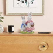 A hand-painted resin figurine of a rabbit family, comprising a mother and child, dressed in decorative outfits and holding a floral basket and a metal pot.