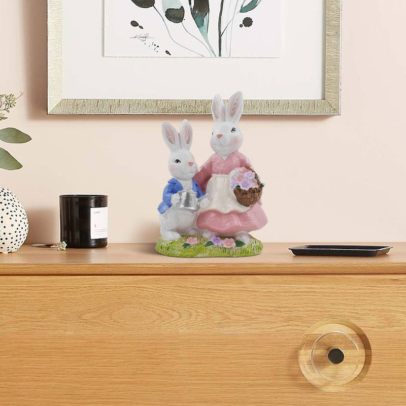 A hand-painted resin figurine of a rabbit family, comprising a mother and child, dressed in decorative outfits and holding a floral basket and a metal pot.